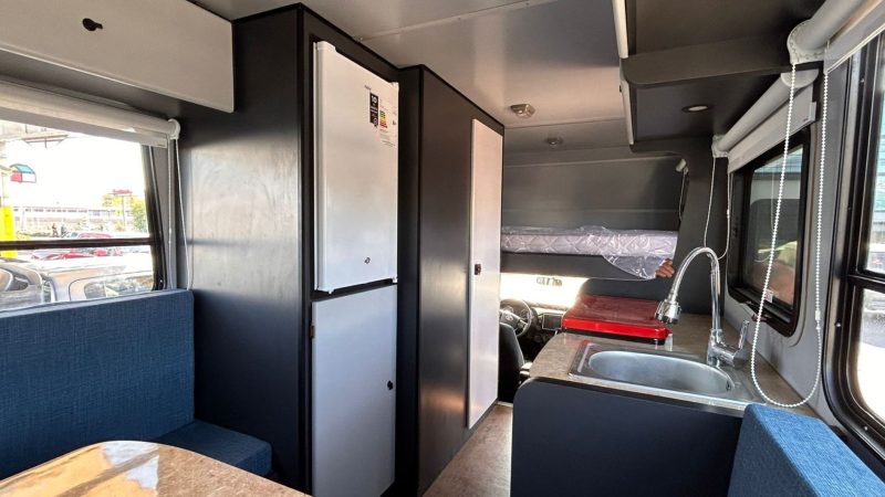 Interior de la camper con refrigerador, cama sobre la cabina, lavadero y cocina integrada, mostrando un uso eficiente del espacio y comodidades modernas.